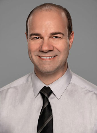 The image shows a man with short hair wearing a dark tie, smiling slightly at the camera. He has a bald spot on top of his head and appears to be indoors with a neutral background.