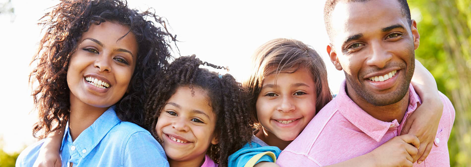 The image shows a family of four with two adults and two children, smiling and posing for a photograph outdoors during daylight.