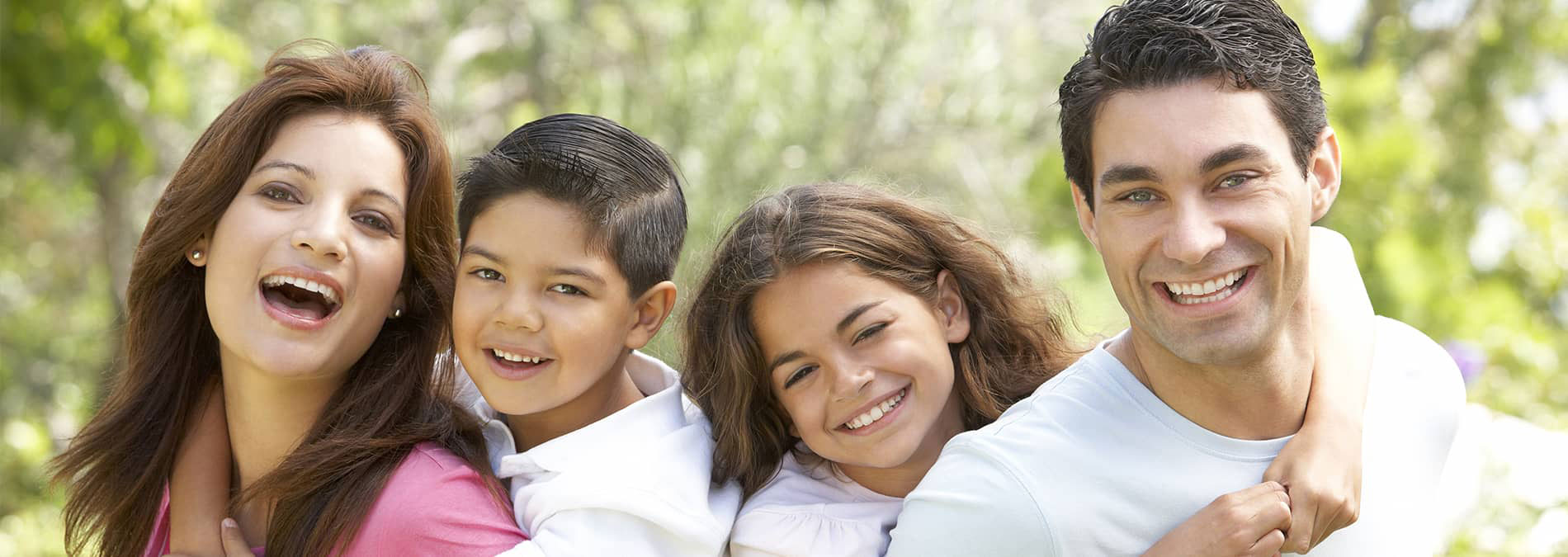 The image shows a family of four with two adults and two children outdoors during daylight, with the adult woman holding one child and smiling at the camera while the other adult male stands behind them, also smiling.