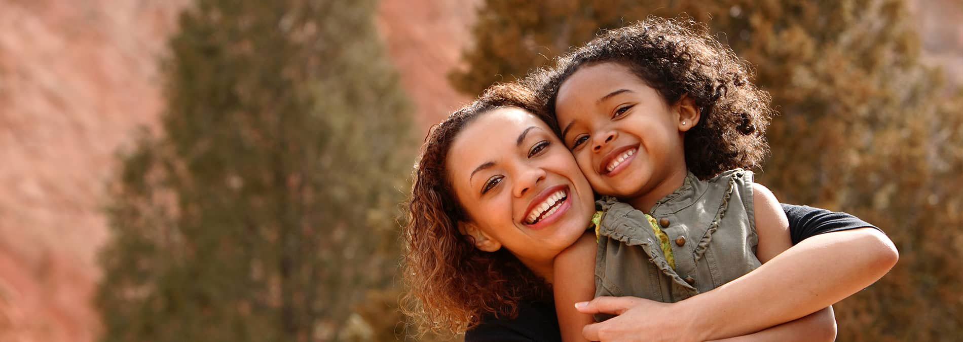 An adult woman and child embrace outdoors during daylight, with a scenic landscape featuring rock formations and greenery in the background.