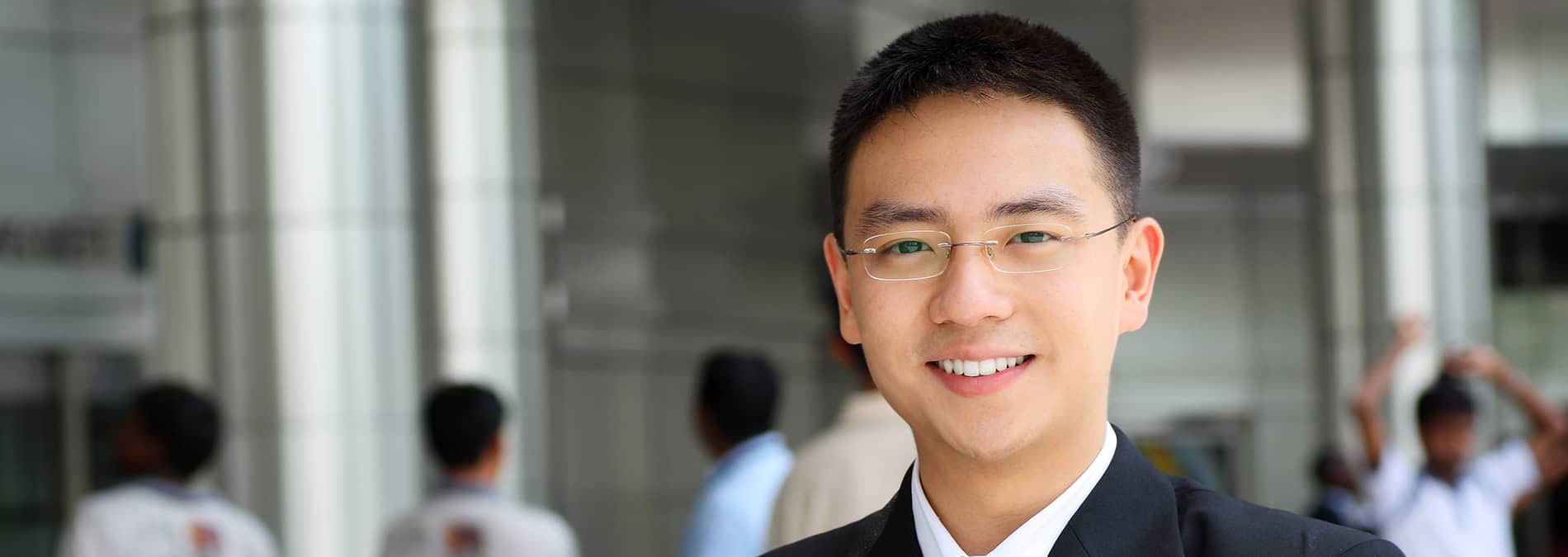This is a photograph of a man standing indoors with a white background behind him. He has short hair, is wearing glasses, and has a beard. His attire includes a dark suit jacket over a light-colored shirt. The man appears to be smiling and looking directly at the camera.