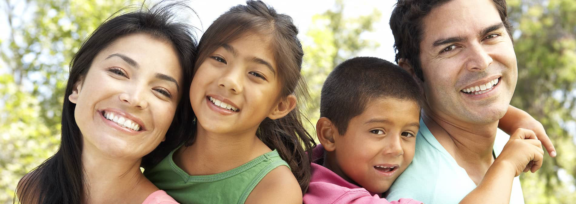 The image shows a family of four with a happy expression, embracing each other outdoors during daylight.