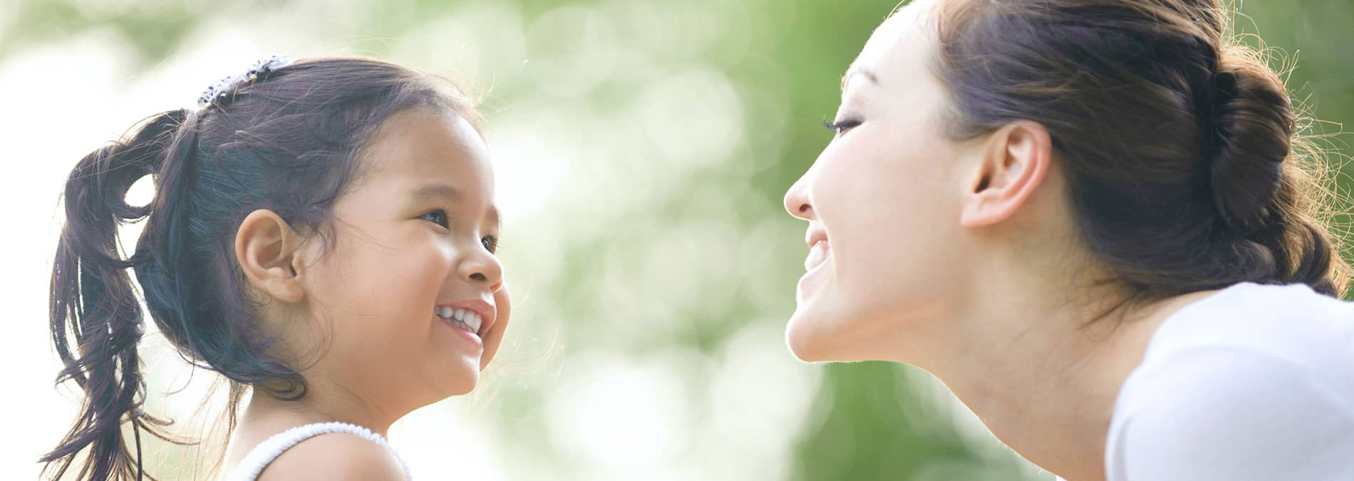 The image shows a smiling woman with her hair up and a child next to her, both looking towards the camera with a blurred background suggesting an outdoor setting.