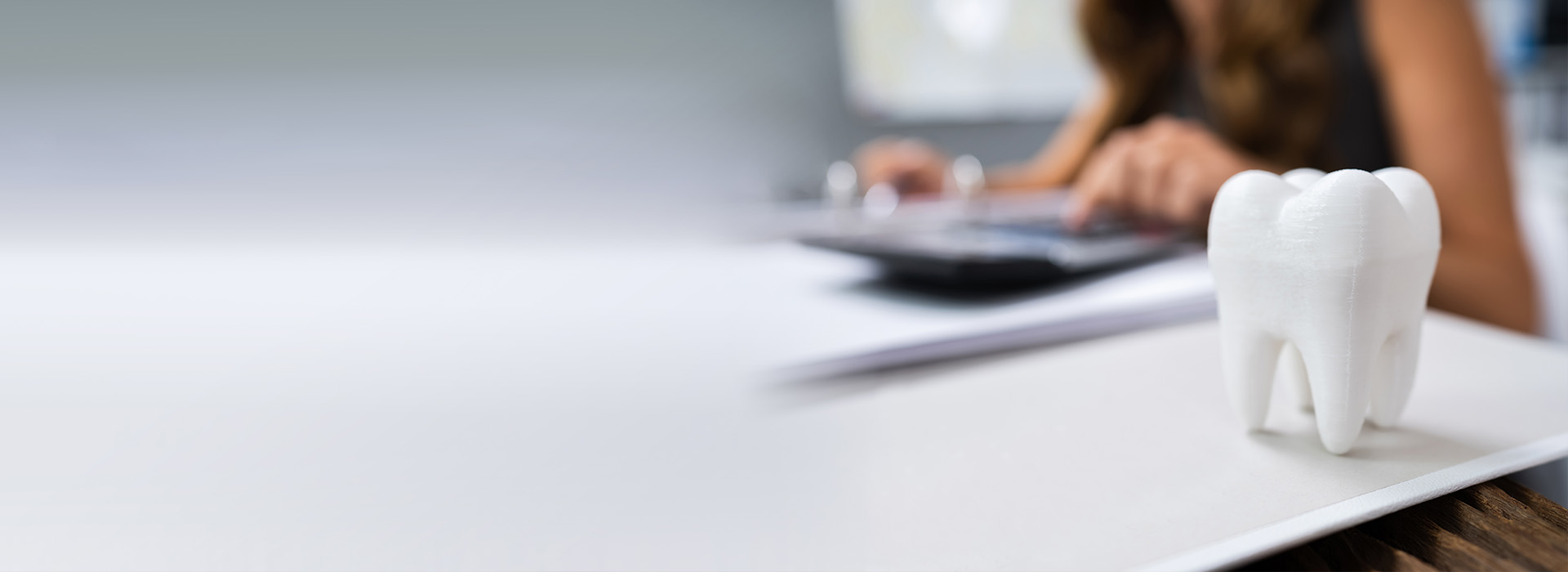 A person sitting at a desk with a computer monitor and keyboard, working on a document, with a 3D model of a tooth placed on top of the paperwork.
