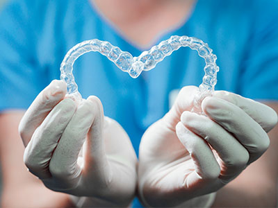 The image shows a pair of clear dental braces being held by hands wearing gloves, with a heart shape formed by the braces  wire.