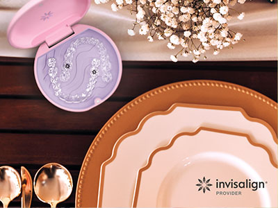 The image shows a table setting with various dinnerware items including plates, bowls, and a spoon rest, along with a pink toothbrush holder with a design on top, placed on a wooden surface with a white tablecloth and a floral arrangement in the background.