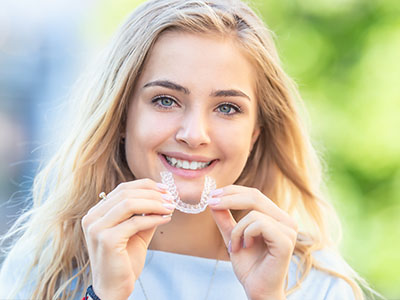 A young woman holding a toothbrush with a smile on her face, set against a blurred outdoor background.