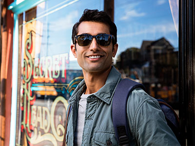 This image features a man standing outdoors, wearing sunglasses, a backpack, and a jacket, with a smile on his face. He is posing for the camera against a backdrop that includes a sign for  Sweet Treats  and a window display.