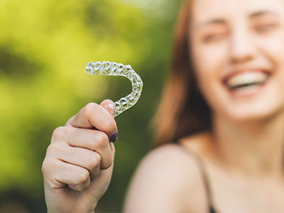 An individual is holding up a clear plastic object, which appears to be a dental retainer, against their smile, with a blurred background of greenery suggesting an outdoor setting.
