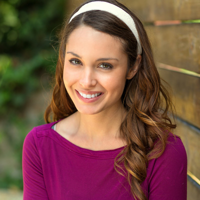 The image shows a young woman with long hair smiling at the camera while wearing a purple top. She has a headband on her head, and she is standing outdoors against a wooden fence.