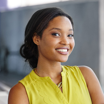 The image shows a person smiling at the camera, wearing a yellow top with a lace-up neckline, against a blurred background that suggests an indoor setting.