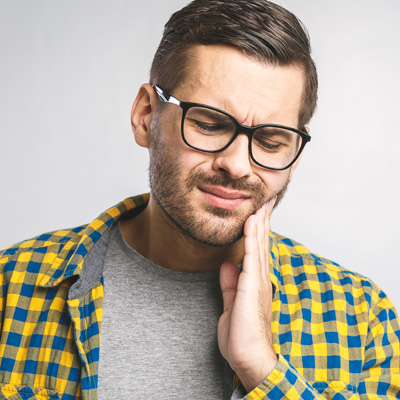 A man with glasses and a beard, possibly in his late 20s or early 30s, wearing a yellow plaid shirt and holding his hand to his mouth as if he s in pain or discomfort.