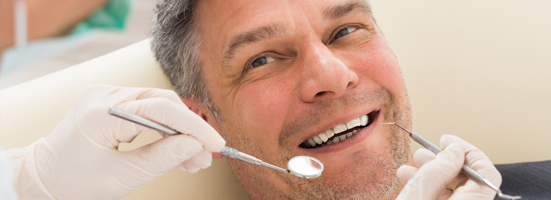 A man sitting in a dental chair with a smiling expression, receiving dental care from a professional wearing gloves and holding tools.