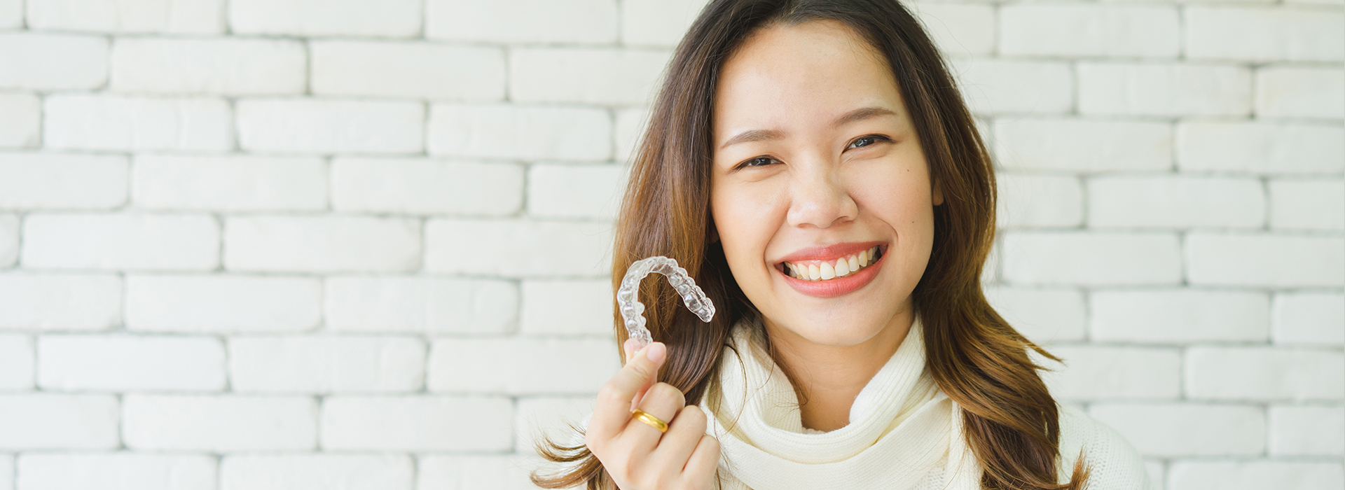 A smiling woman wearing a ring holds a small object with a round top, set against a brick wall background.