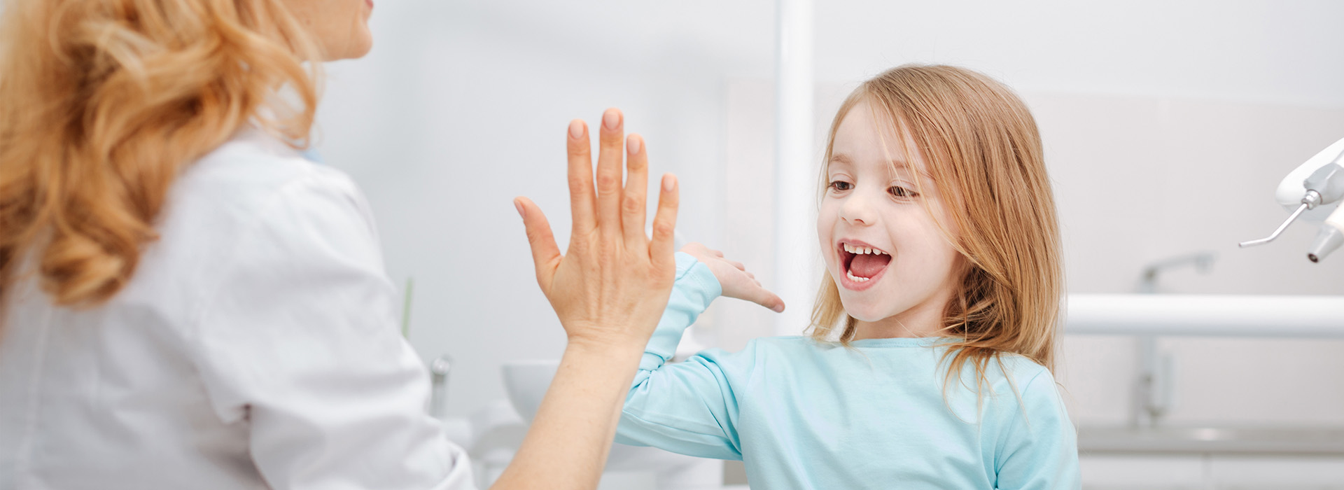 This is a photograph featuring an adult woman and a young girl with their hands together, smiling at each other. The setting appears to be a dental office, indicated by the presence of dental equipment and a sink in the background.
