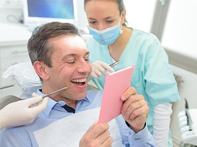 A man sitting in a dental chair, holding a pink card with a smile on his face, while a female dentist stands nearby, smiling and looking at him.