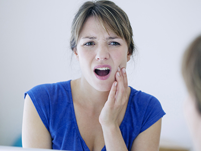 A woman with her mouth open, looking at her reflection in a mirror while standing in front of a sink.