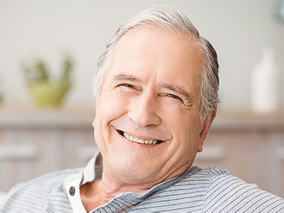 The image shows an elderly man with white hair, smiling at the camera while sitting comfortably in a chair indoors.